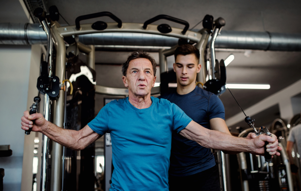 A senior man with a young male trainer doing strength workout exercise in gym.