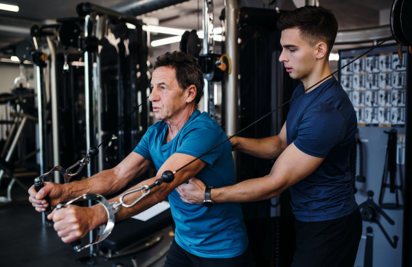 A senior man with a young male trainer doing strength workout exercise in gym.