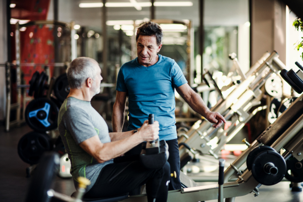 A senior man with a young male trainer doing strength workout exercise in gym.