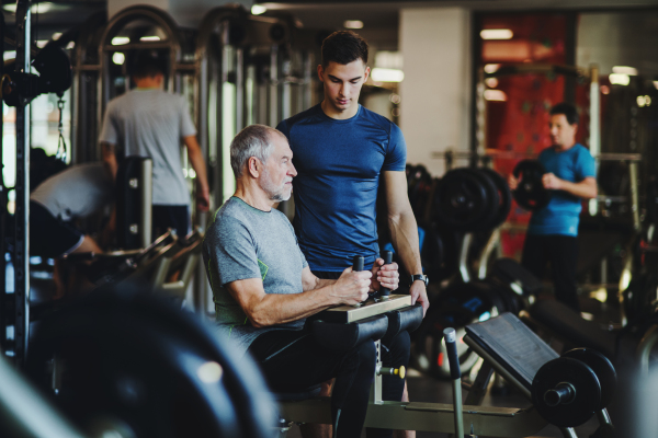 A senior man with a young male trainer doing strength workout exercise in gym.