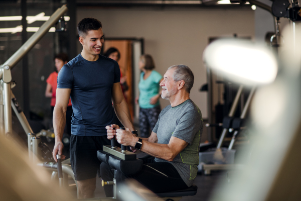 A senior man with a young male trainer doing strength workout exercise in gym.