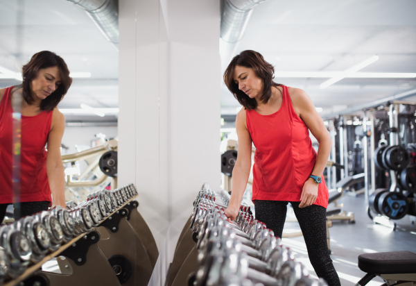 A female senior in gym doing exercise with dumbbells, a reflection in mirror.