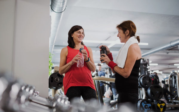 Two cheerful senior women in gym resting after doing exercise, holding water bottles.