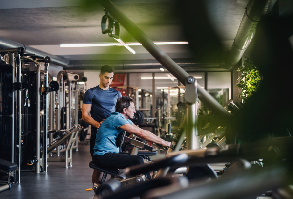 A senior man with a young male trainer doing strength workout exercise in gym.