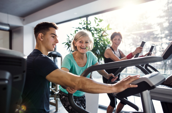 Two cheerful female seniors in gym with a young trainer doing cardio work out, exercising on stationary bicycle.