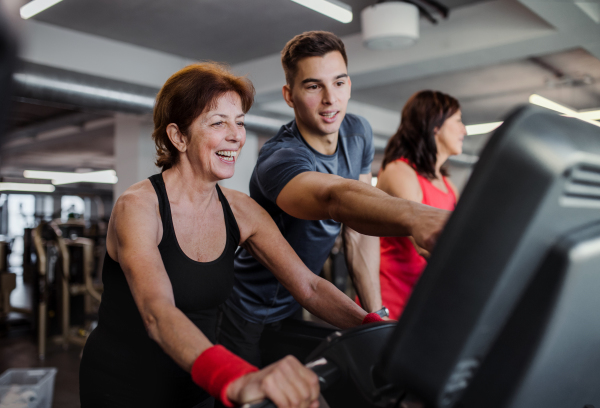 Two cheerful female seniors in gym with a young trainer doing cardio work out, exercising on stationary bicycle.