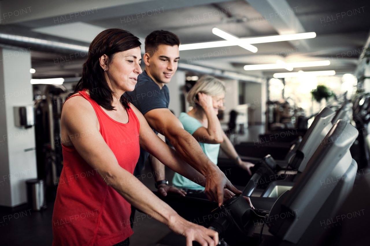 Two cheerful female seniors in gym with a young trainer doing cardio work out, exercising on stationary bicycle.