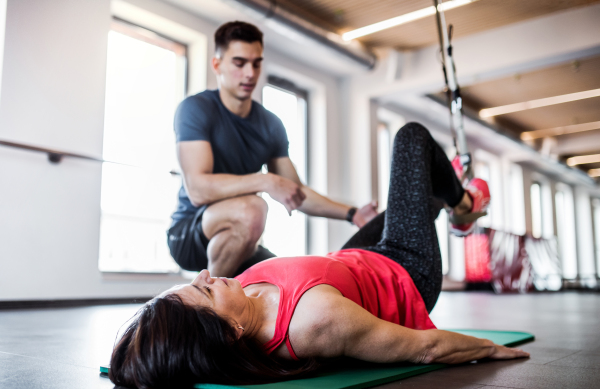 A senior woman in gym and a handsome personal trainer doing exercise with TRX.