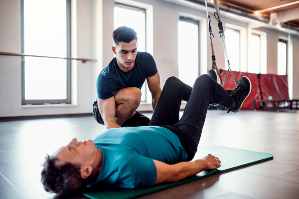 A senior man in gym and a handsome personal trainer doing exercise with TRX.