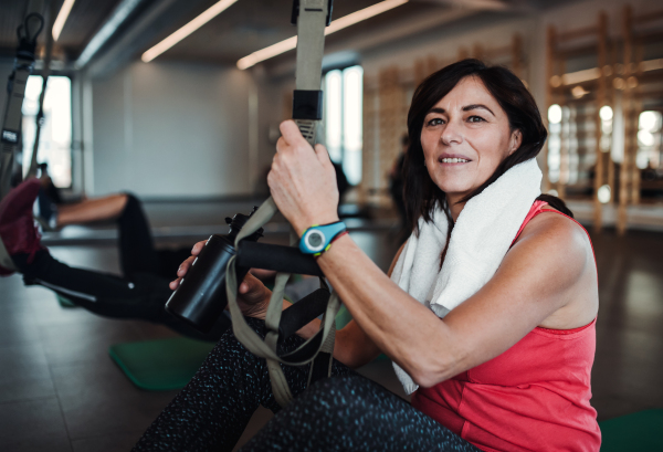 A portrait of happy senior woman in gym doing exercise with TRX. Copy space.