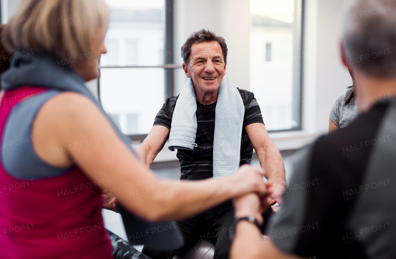 A group of cheerful seniors in gym resting after doing exercise on fit balls, holding hands and talking.