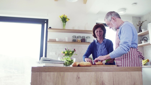 A portrait of happy senior couple in love indoors at home, preparing food.