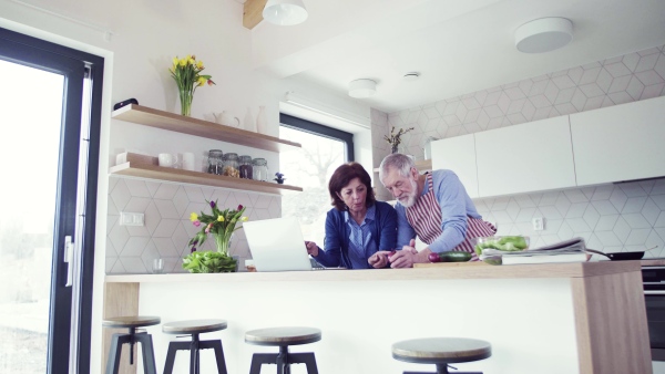 A portrait of happy senior couple with laptop indoors at home, cooking.