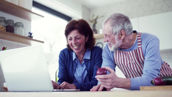 A portrait of happy senior couple with laptop indoors at home, cooking.