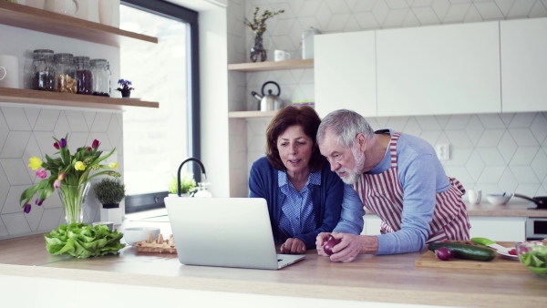 A portrait of happy senior couple with laptop indoors at home, cooking.