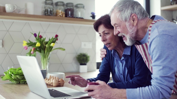 A portrait of happy senior couple with laptop indoors at home, cooking.