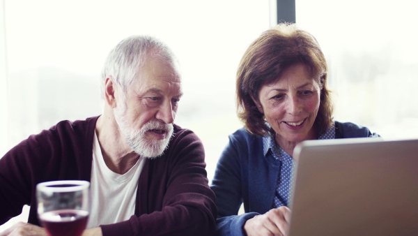 A portrait of happy senior couple with wine indoors at home, using laptop.