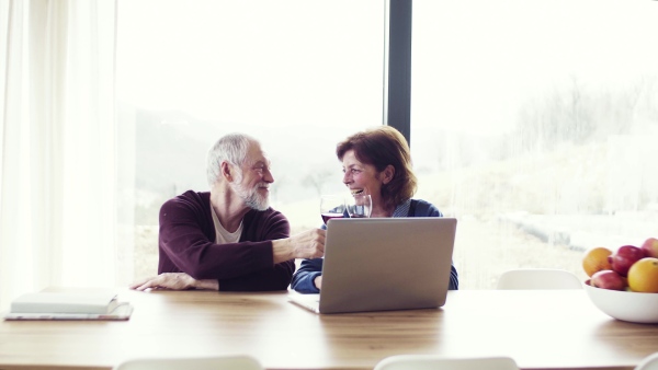 A portrait of happy senior couple with wine indoors at home, using laptop.