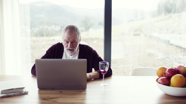 A portrait of happy senior man with wine indoors at home, using laptop.