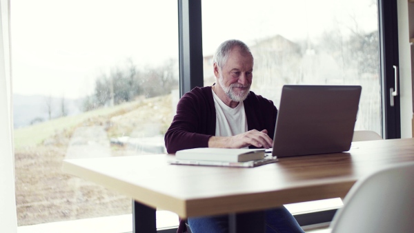 A portrait of happy senior man indoors at home, using laptop.