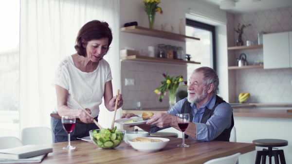 A senior couple in love serving lunch indoors at home, talking.