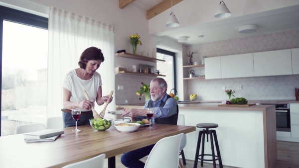 A senior couple in love serving lunch indoors at home, talking.