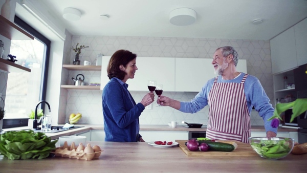 A portrait of happy senior couple in kitchen indoors at home, clinking glasses when cooking. Slow motion.