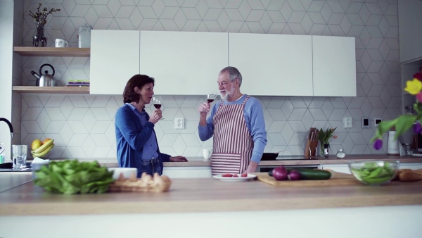 A portrait of happy senior couple in kitchen indoors at home, clinking glasses when cooking. Slow motion.