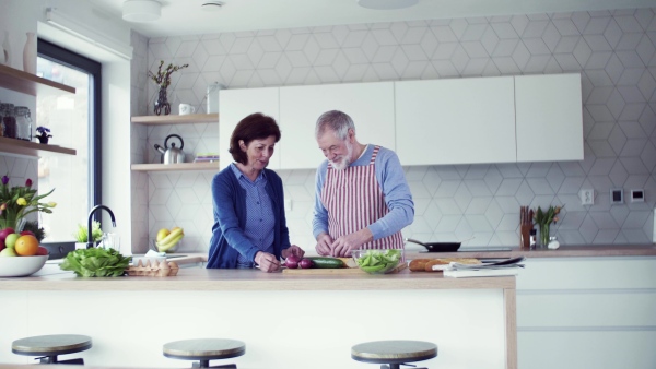 A portrait of happy senior couple in love indoors at home, preparing food.