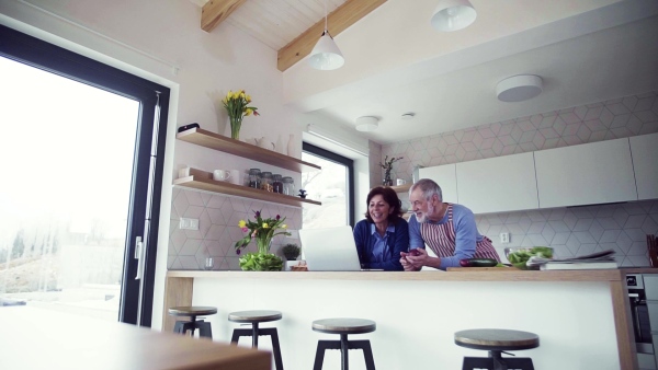 A portrait of happy senior couple with laptop indoors at home, cooking. Slow motion.