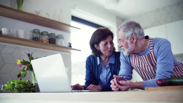 A portrait of happy senior couple with laptop indoors at home, cooking. Slow motion.