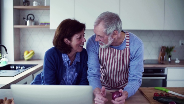 A portrait of happy senior couple with laptop indoors at home, cooking. Slow motion.