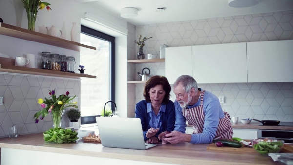 A portrait of happy senior couple with laptop indoors at home, cooking. Slow motion.