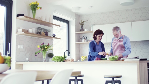 A portrait of happy senior couple in love indoors at home, preparing food.