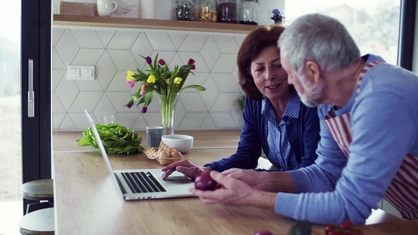 A portrait of happy senior couple with laptop indoors at home, cooking. Slow motion.