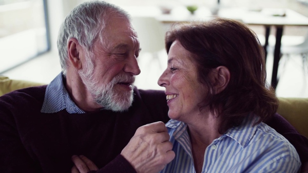 An affectionate senior couple in love sitting on sofa indoors at home, talking and kissing. Slow motion.