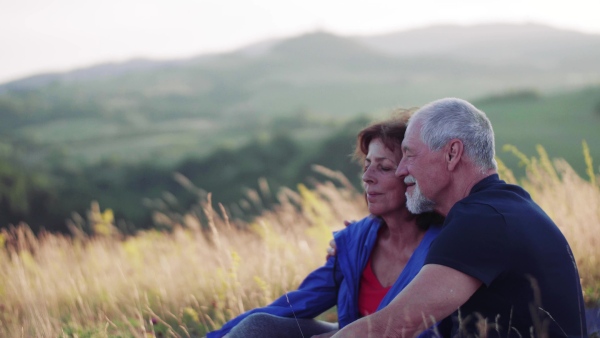 A senior tourist couple in love sitting in nature at sunset, resting.