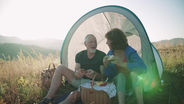 A senior tourist couple with shelter sitting in nature at sunset, eating sandwiches. Slow motion.