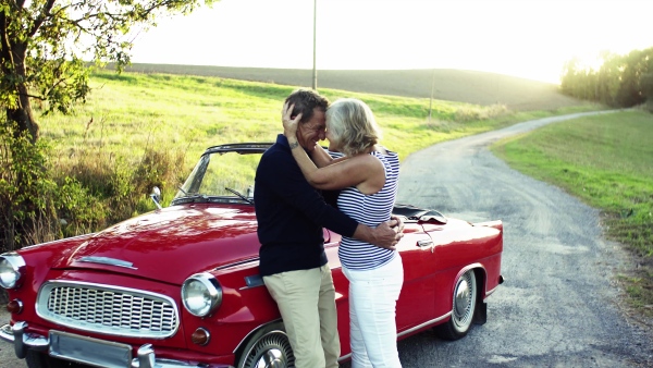 A cheerful senior couple in love standing by cabriolet on a road trip in summer at sunset, hugging.