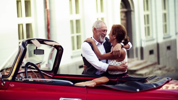 A cheerful senior couple in love standing by cabriolet on a road trip in summer in town, hugging. Slow motion.