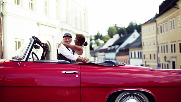 A cheerful senior couple in love standing by cabriolet on a road trip in summer in town at sunset, hugging. Slow motion.