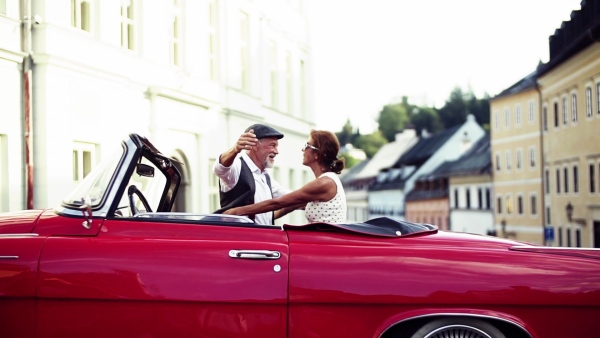 A cheerful senior couple in love standing by cabriolet on a road trip in summer in town, hugging and kissing. Slow motion.