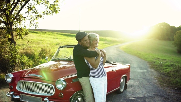 A cheerful senior couple in love standing by cabriolet on a road trip in summer at sunset. Slow motion.