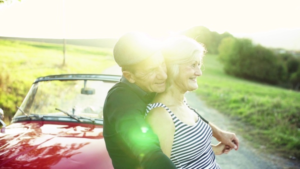 A cheerful senior couple in love standing by cabriolet on a road trip in summer at sunset. Slow motion.