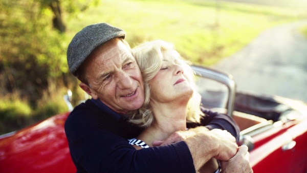 A cheerful senior couple in love standing by cabriolet on a road trip in summer at sunset. Slow motion.