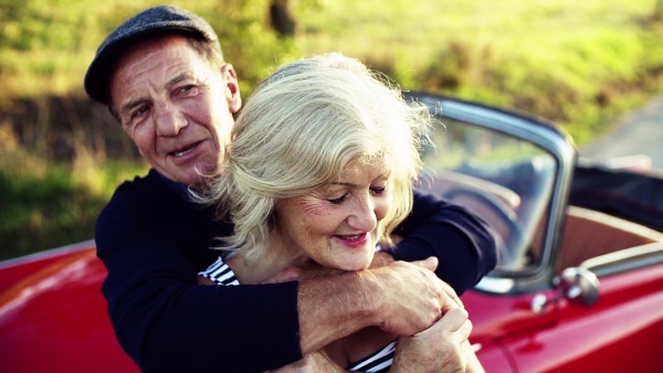 A cheerful senior couple in love standing by cabriolet on a road trip in summer at sunset, hugging. Slow motion.