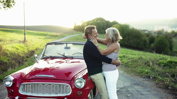 A cheerful senior couple in love standing by cabriolet on a road trip in summer at sunset. Slow motion.