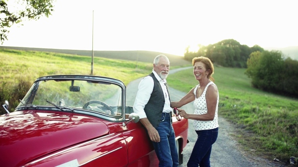 A cheerful senior couple in love standing by cabriolet on a road trip in summer at sunset, talking. Slow motion.