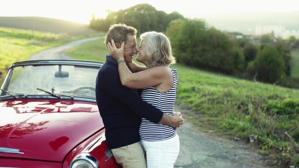 A cheerful senior couple in love standing by cabriolet on a road trip in summer at sunset, hugging.