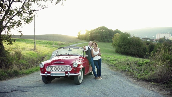 A cheerful senior couple in love standing by cabriolet on a road trip in summer at sunset, talking. Slow motion.